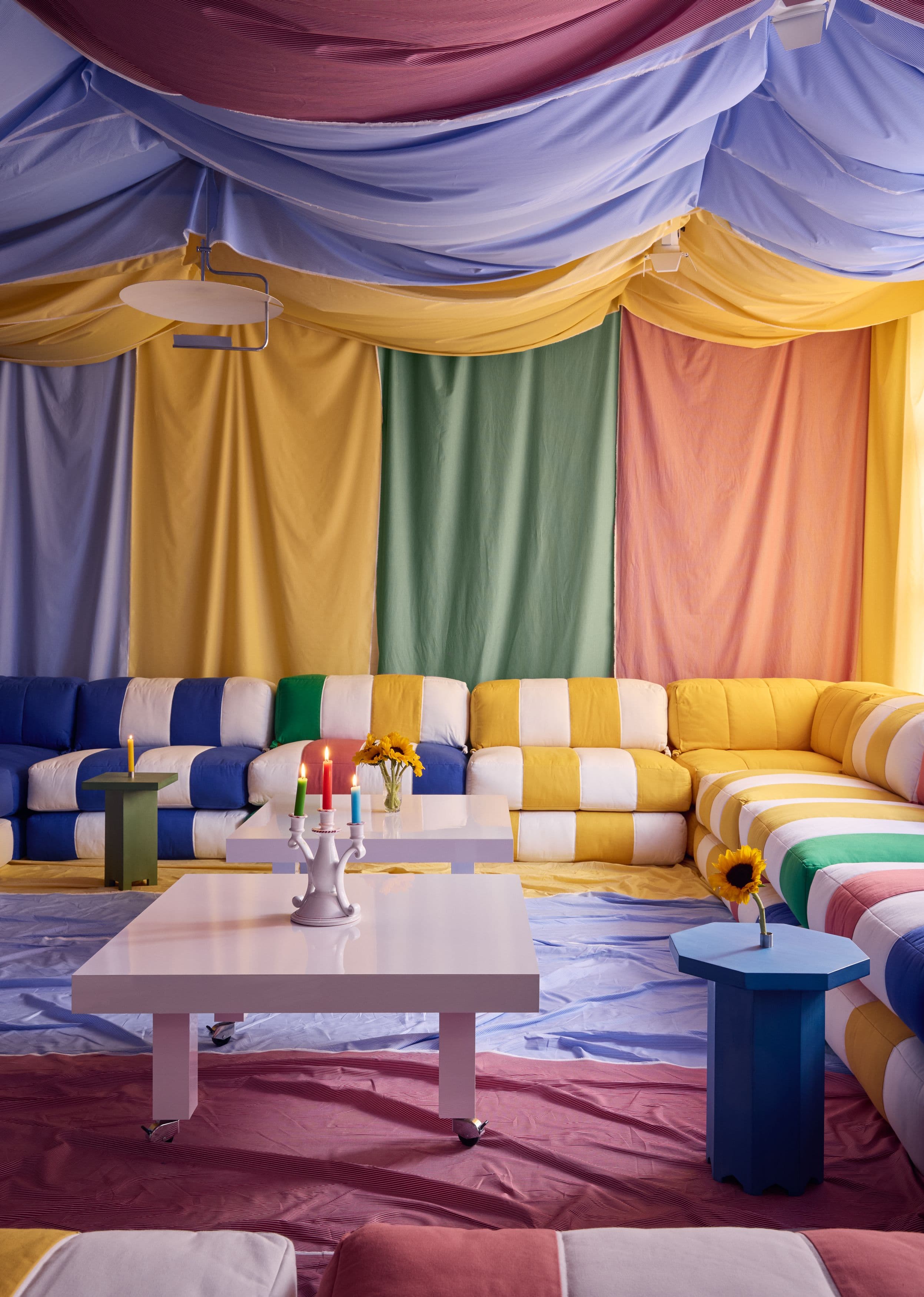 Blue, yellow and multicolored striped seating beneath fabric-draped walls and ceiling, with pink tables and a blue side table
